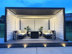 Contemporary outdoor lounge with a black louvered pergola, integrated LED lighting, cushioned seating, and potted plants on a wooden deck at dusk