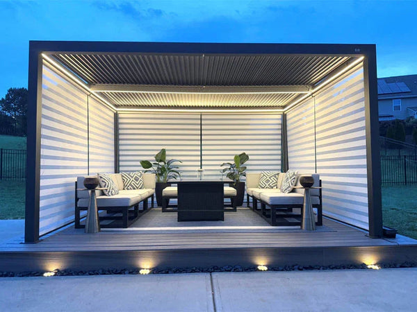 Contemporary outdoor lounge with a black louvered pergola, integrated LED lighting, cushioned seating, and potted plants on a wooden deck at dusk