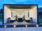 Contemporary outdoor lounge with a black louvered pergola, integrated LED lighting, cushioned seating, and potted plants on a wooden deck at dusk