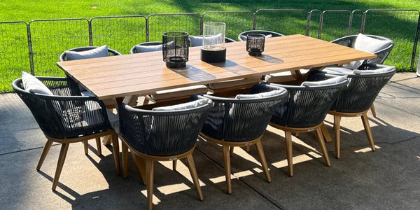 Modern outdoor dining set with a long wooden table and eight rope-woven chairs with gray cushions on a patio, surrounded by green grass