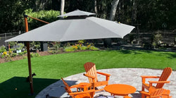Large white cantilever patio umbrella shading four orange Adirondack chairs and a round orange table on a circular stone patio in a backyard garden