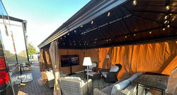 Outdoor gazebo with string lights, wicker furniture, a TV, and floor lamps, set up on a brick patio next to an RV