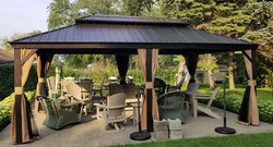 Rectangular hardtop gazebo with black metal frame and brown curtains, containing various outdoor seating including Adirondack chairs and wicker furniture on a concrete patio surrounded by manicured lawn