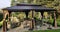 Rectangular hardtop gazebo with black metal frame and brown curtains, containing various outdoor seating including Adirondack chairs and wicker furniture on a concrete patio surrounded by manicured lawn