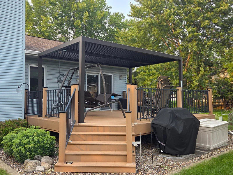 A single-story white brick house with a black pergola covering the patio area. The landscaped yard features shrubs, flowers, and stepping stones across a well-maintained lawn