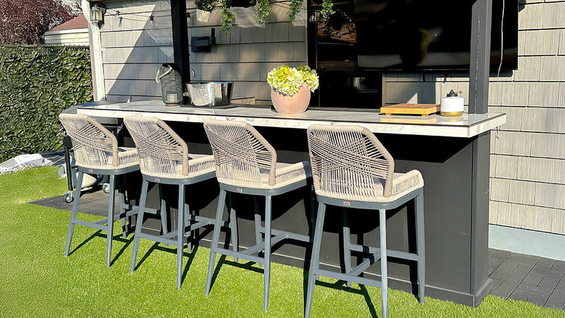 Modern outdoor bar setup with four rope-woven bar stools in beige/gray color against a gray exterior wall. The stools feature contemporary design with woven seats and backs, metal frames, and are placed at a sleek countertop with decorative potted plants