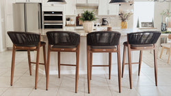 A modern kitchen featuring four stylish bar stools with wooden frames and woven backs positioned at a white island countertop, complemented by stainless steel appliances and white cabinetry