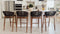 A modern kitchen featuring four stylish bar stools with wooden frames and woven backs positioned at a white island countertop, complemented by stainless steel appliances and white cabinetry