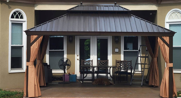 A brown metal gazebo with tan curtains provides shade for a patio dining set. A golden retriever dog relaxes on the concrete patio