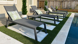 Row of modern gray outdoor chaise lounges with green leaf-patterned pillows arranged alongside a swimming pool, set on artificial grass strips with small side tables between them
