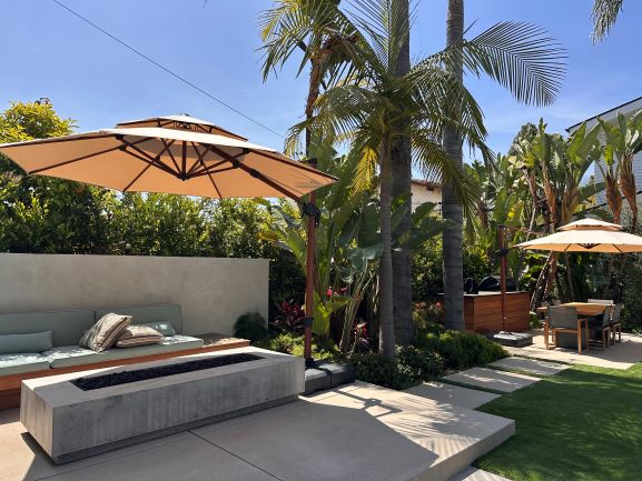 Modern backyard oasis featuring a beige cantilever umbrella shading a concrete bench seating area with built-in fire pit, surrounded by palm trees and tropical plants, with artificial turf lawn and additional seating area with umbrella in the background