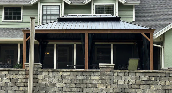 Modern gazebo with black metal roof and wooden posts attached to a house, built on a stone wall patio foundation