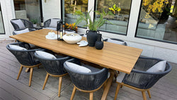 A stylish outdoor dining setup featuring a wooden table surrounded by modern rope-woven chairs with cushions, placed on a deck in front of a house with large windows