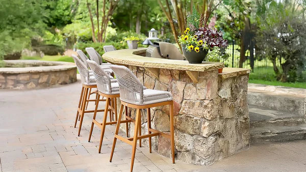 Rustic stone outdoor bar counter with rough-cut stone finish, featuring four woven bar stools with wooden frames and light cushions, overlooking a garden landscape with decorative flower pot on the countertop