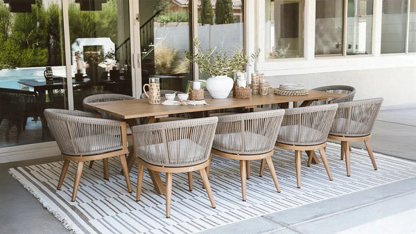 Outdoor dining setup with a long wooden table and woven chairs, surrounded by greenery and decorated with various tableware