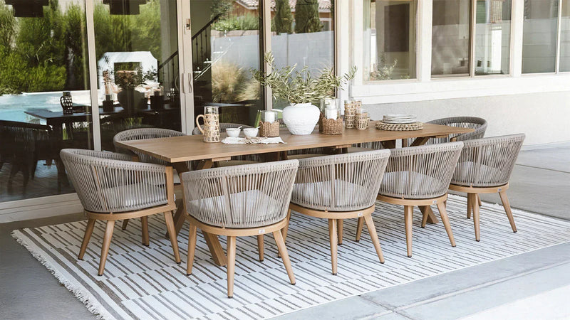 Outdoor dining setup with a long wooden table and woven chairs, surrounded by greenery and decorated with various tableware