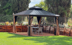 Elegant black metal hardtop gazebo with tan curtains in a well-maintained garden. The structure sits on a circular gravel base surrounded by green lawn and wooden raised garden beds