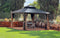 Elegant black metal hardtop gazebo with tan curtains in a well-maintained garden. The structure sits on a circular gravel base surrounded by green lawn and wooden raised garden beds