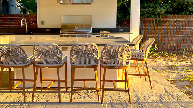 An outdoor kitchen with a counter and six woven chairs, set against a brick wall.