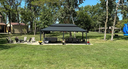 Modern black double-tiered gazebo with brown curtains set on a concrete patio, furnished with various outdoor chairs and seating arrangements in a landscaped backyard