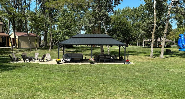 Modern black double-tiered gazebo with brown curtains set on a concrete patio, furnished with various outdoor chairs and seating arrangements in a landscaped backyard