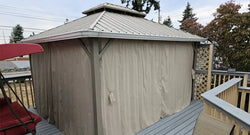 Outdoor metal gazebo with beige privacy curtains on a wooden deck, surrounded by trees and outdoor furniture