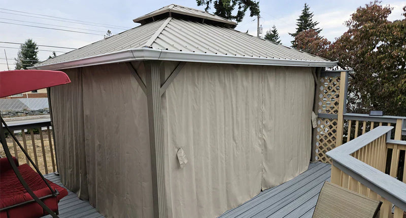 Outdoor metal gazebo with beige privacy curtains on a wooden deck, surrounded by trees and outdoor furniture